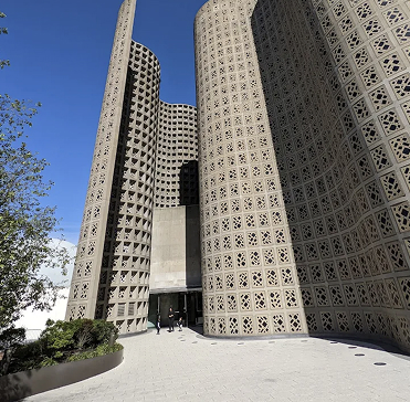 Tall modern building with rounded, perforated concrete walls and geometric patterns, an entrance at the center, and people walking nearby under a clear blue sky.
